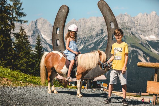Going_PonyAlm_Wilder Kaiser Zwei Kinder stehen neben einem Ponny in den Bergen. Im Hintergrund sind hohe Berge und ein blauer Himmel zu sehen.