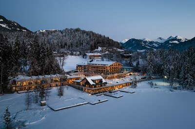 Alpenhotel Kitzbühel im Winter Beleuchtetes Hotel am verschneiten Seeufer vor bewaldeten Bergen in der Dämmerung.