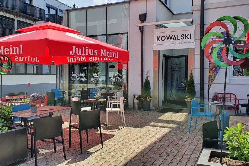 Cafe Kowalski Ein einladendes Café mit roten Sonnenschirmen und bunten Stühlen. Die Terrasse ist hell und freundlich mit Pflanzen und einem Spielrad.