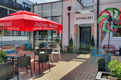 Cafe Kowalski Ein einladendes Café mit roten Sonnenschirmen und bunten Stühlen. Die Terrasse ist hell und freundlich mit Pflanzen und einem Spielrad.