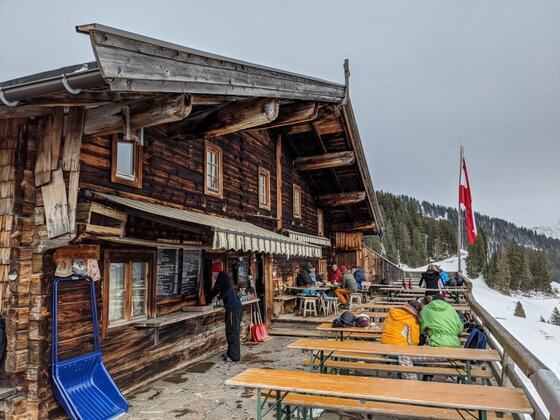Terrasse der Trattenbachalm Rustikale Holzhütte mit bewirtschafteter Terrasse, Gästen an Holztischen und einer rot-weißen Flagge im Schnee.