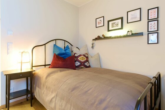 Schlafbereich im Helene Apartment Einzelbett mit Metallrahmen, bunten Kissen, Nachttisch und Wanddekoration in einem hellen Zimmer.