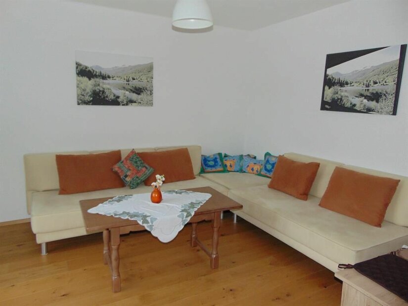 Wohnzimmer mit Ecksofa, Holztisch und Landschaftsbildern an weißen Wänden.