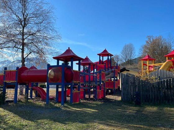 Großes rotes Spielgerüst mit Türmen, Rutschen und Röhren auf einer Wiese vor Bergen.