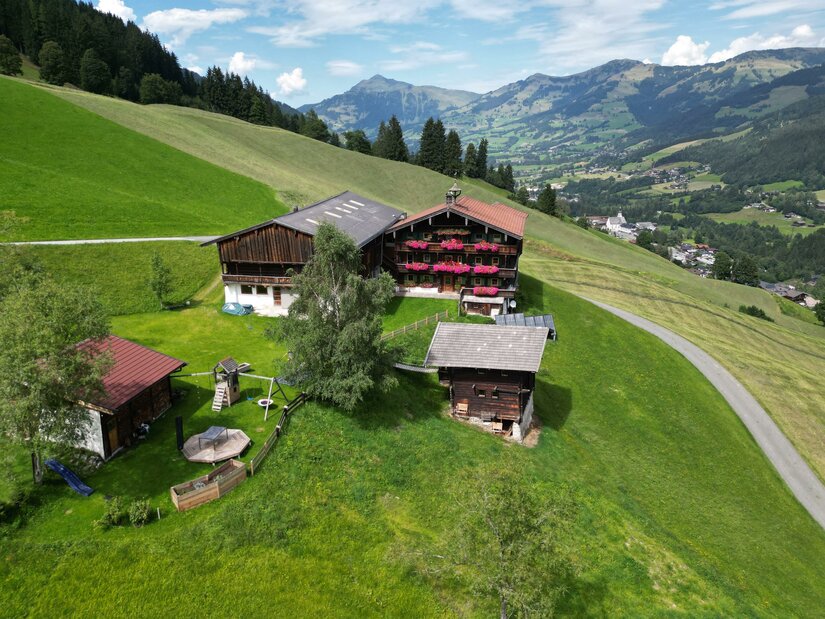 THENN im Sommer Luftaufnahme eines traditionellen Holzbauernhofs mit Blumenschmuck auf einer grünen Bergwiese.