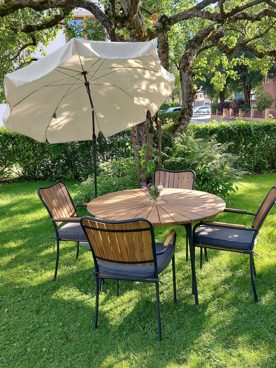 Gartenwohnung Ritter Außenbereich Runder Holztisch mit vier Stühlen und einem Sonnenschirm auf einer Rasenfläche unter einem Baum.