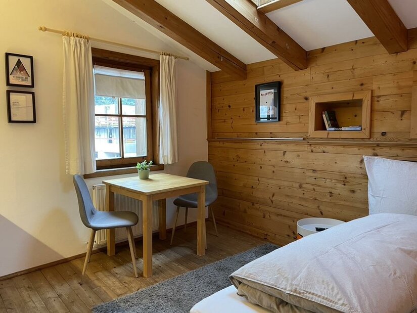 Zimmer mit Holzwänden, Bett, Holztisch, zwei grauen Stühlen und einem Fenster.
