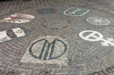 Eine dekorative Pflasterung mit verschiedenen Wappen und Symbolen. Die Gestaltung zeigt kunstvolle Muster auf dem Boden.
