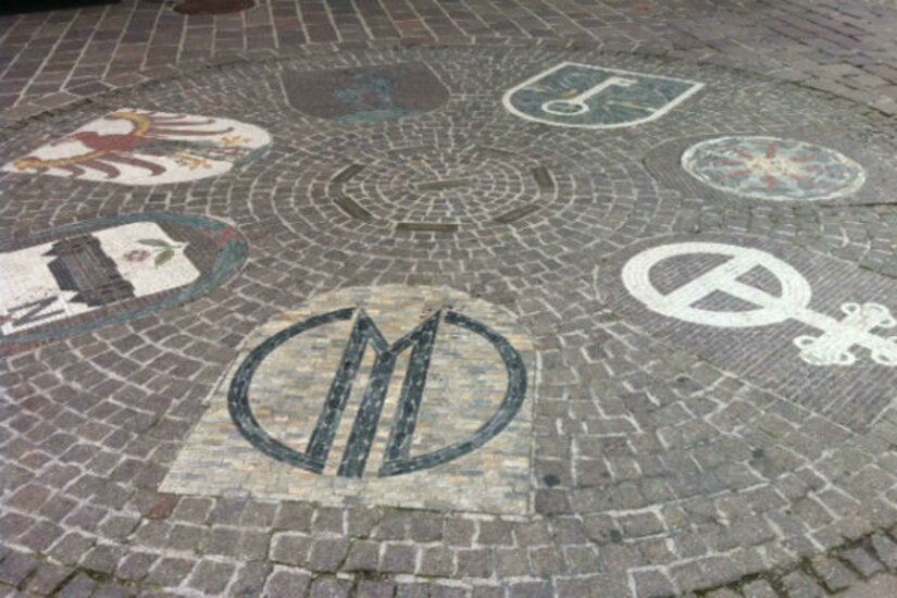 Stadtwappen der Schwesternstädte Eine dekorative Pflasterung mit verschiedenen Wappen und Symbolen. Die Gestaltung zeigt kunstvolle Muster auf dem Boden.