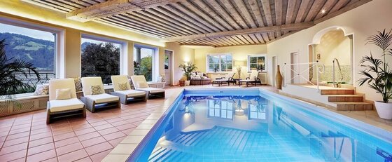 Spa de Charme Tennerhof Hallenbad mit Liegestühlen, Holzdecke und großen Fenstern mit Blick auf die Berge.