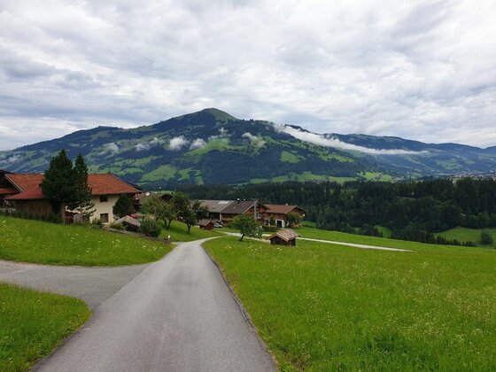 Wanderweg bei Lend-Kinzlingalm Asphaltierter Weg führt durch grüne Wiesen zu einer kleinen Siedlung vor bewaldeten Bergen unter bewölktem Himmel.