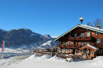 Berggasthof Hagstein im Winter Rustikales Holzhaus mit Terrasse in einer verschneiten Berglandschaft unter blauem Himmel.