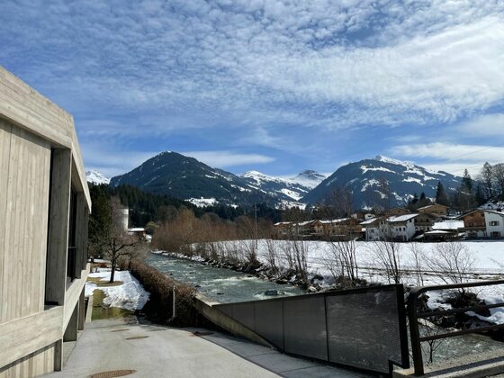 Achenweg Aurach - Jochberg Blick auf einen Flusslauf neben einem modernen Gebäude mit schneebedeckten Bergen im Hintergrund.