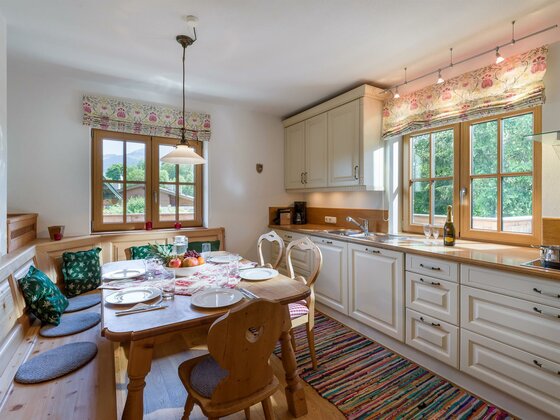 Wohnküche Appartement Hahnenkamm Helle Landhausküche mit Holztisch, Eckbank und weißer Küchenzeile vor zwei Fenstern.