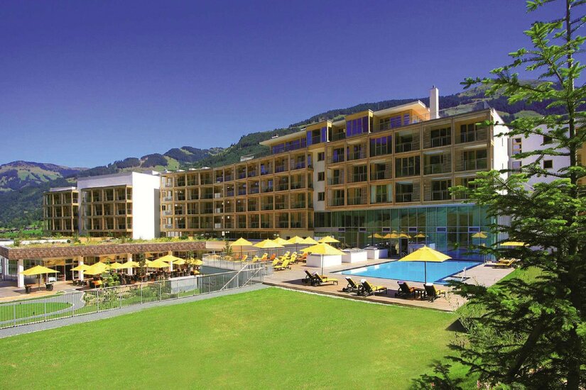 Kempinski Hotel im Sommer Modernes Hotelgebäude mit Außenpool, gelben Sonnenschirmen und Liegewiese vor bewaldeten Bergen.