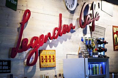 Roter Schriftzug Legenden Cafe an einer Holzwand mit Getränkeflaschen und Schildern.