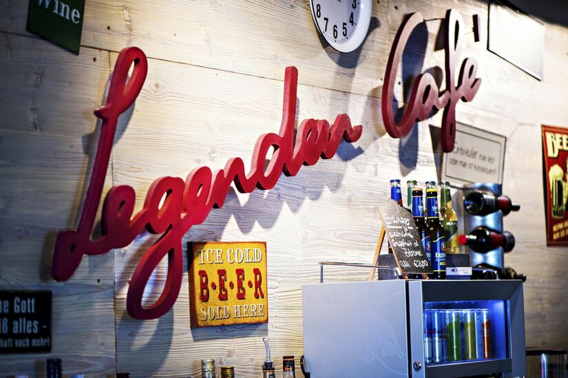 Legenden Cafe in Kitzbühel Roter Schriftzug Legenden Cafe an einer Holzwand mit Getränkeflaschen und Schildern.