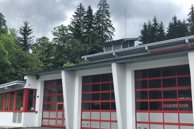 Feuerwehr <p>Ein modernes Feuerwehrgebäude mit roten Garagentoren in Aurach. Im Hintergrund sind hohe Bäume und graue Wolken zu sehen.</p>