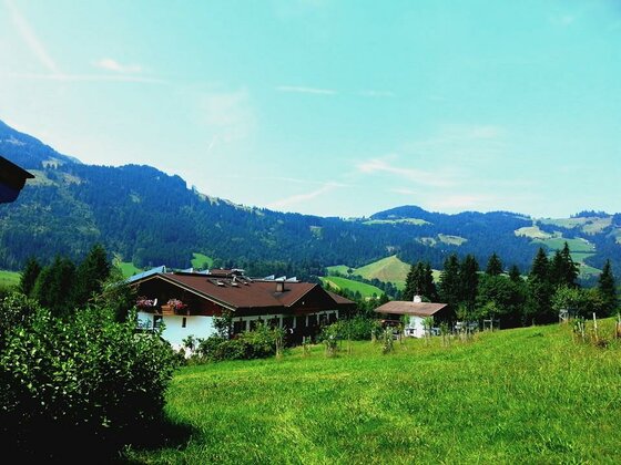 Alpines Gebäude mit Satteldach auf einer grünen Wiese vor bewaldeten Bergen.