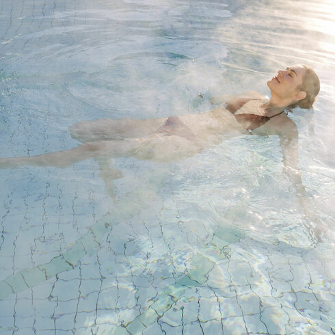 Eine Frau entspannt im warmen Wasser eines Spas, umgeben von Dampf und sanften Lichtreflexionen auf der Wasseroberfläche.