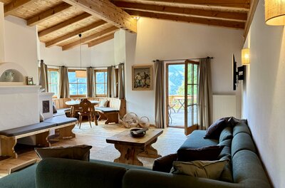 Ein einladendes Wohnzimmer mit Holzbalkendecke und großen Fenstern. Gemütliche Sitzgelegenheiten und ein Esstisch schaffen eine warme Atmosphäre.