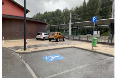 Behindertenparkplatz - ÖBB Hauptbahnhof Kitzbühel Ein Parkplatz mit zwei Autos, darunter ein behindertengerechter Parkplatz. Im Hintergrund ist eine Bahnhofsplattform und Bäume zu sehen.