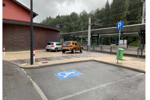 Ein Parkplatz mit zwei Autos, darunter ein behindertengerechter Parkplatz. Im Hintergrund ist eine Bahnhofsplattform und Bäume zu sehen.