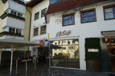 Ein McCafé an einem Gebäudefront mit großen Fenstern. Draußen stehen ein paar Tische und Sonnenschirme.
