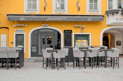 Bar Jimmy's Außenansicht Außenansicht der Bar Jimmy's mit gelber Fassade, grauer Markise und Stehtischen im Außenbereich.