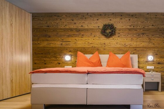 Schlafzimmer im Hoamatl Jochberg Doppelbett mit orangefarbener Bettwäsche vor einer rustikalen Holzwand mit Kranz.
