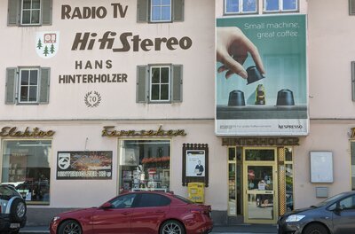 Hinterholzer Ein Geschäft mit einem großen Schild für HiFi-Geräte und Elektrotechnik. Nebenan ist eine Werbung für Nespresso-Kaffeemaschinen.