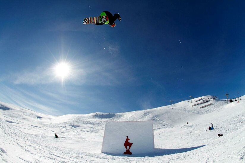 KitzSki Snowpark Hanglalm Ein Snowboarder springt über ein großes Hindernis im Schnee, während die Sonne hell am Himmel scheint. Die Landschaft ist schneebedeckt und die Umgebung ist klar und sonnig.