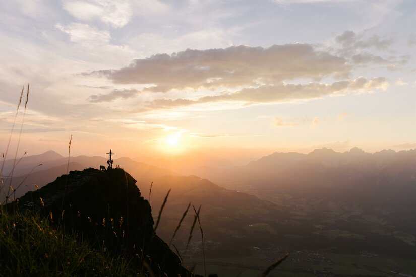 Silhouette eines Gipfelkreuzes auf einem Felsvorsprung vor einer Bergkulisse bei Sonnenuntergang.