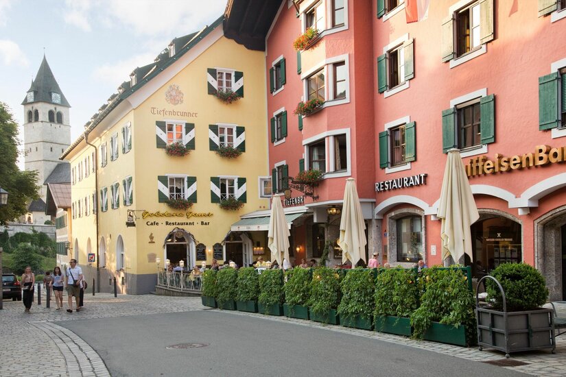 Hotel Tiefenbrunner in Kitzbühel Blick auf farbige Häuserfassaden mit Restaurants und einem Kirchturm im Hintergrund in einer gepflasterten Straße.