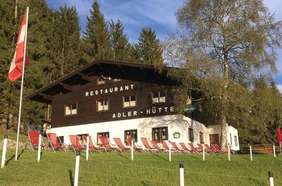 Adlerhütte im Sommer Zweistöckiges Holzgebäude mit weißem Erdgeschoss, roten Liegestühlen davor und einer österreichischen Flagge.