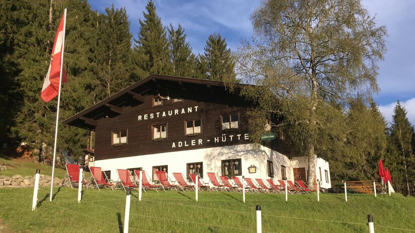 Zweistöckiges Holzgebäude mit weißem Erdgeschoss, roten Liegestühlen davor und einer österreichischen Flagge.