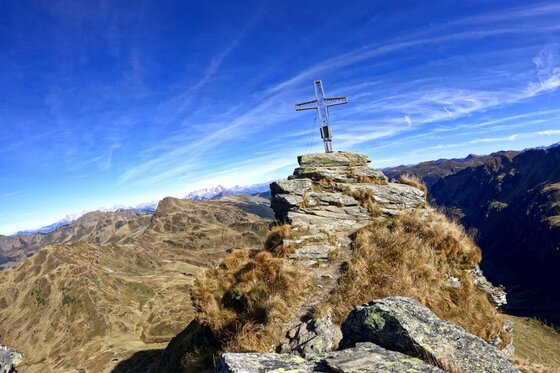 Gipfelkreuz am Tristkogel Ein metallisches Gipfelkreuz auf einem felsigen Berggipfel vor blauem Himmel und Gebirgspanorama.