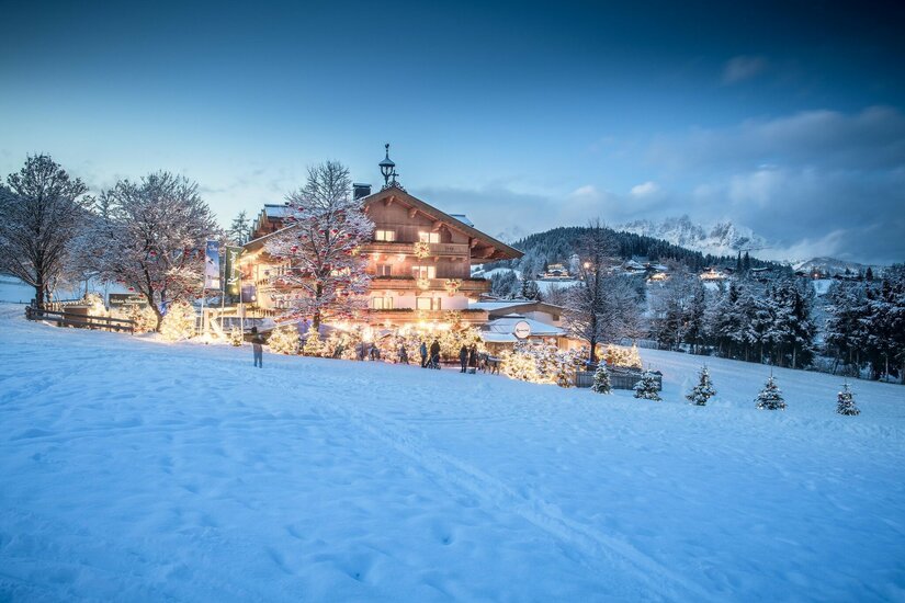 Beleuchtetes Holzhotel in verschneiter Landschaft bei Dämmerung mit geschmückten Tannenbäumen.