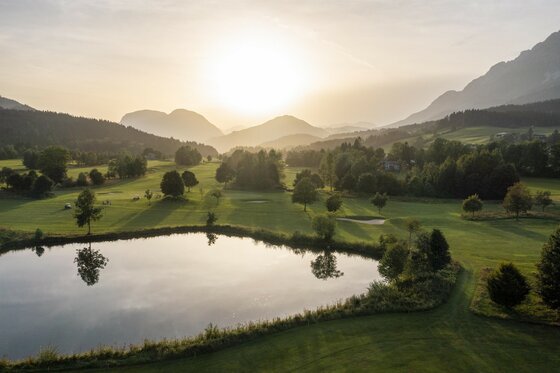 Eine malerische Landschaft mit sanften Hügeln und einem ruhigen Teich. Die Sonne geht hinter den Bergen unter und taucht die Szene in warmes Licht.