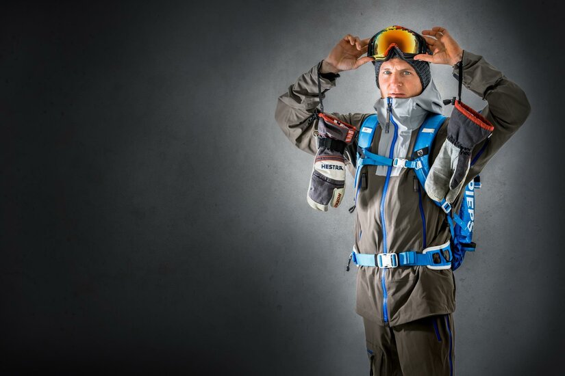 Ein Mann trägt eine Skiausrüstung und hält eine Skibrille in den Händen. Er steht vor einem grauen Hintergrund und wirkt bereit für ein Abenteuer im Schnee.