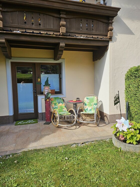 Zwei Korbsessel mit grünen Polstern und ein kleiner Tisch auf einer gepflasterten Terrasse unter einem Holzbalkon.