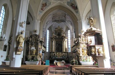 Innenansicht einer barocken Kirche mit goldenem Hochaltar, Kanzel, Kirchenbänken und Deckenmalereien.