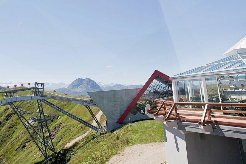 Moderne Bergstation mit Glaspavillon und Seilbahnstütze vor alpinem Panorama.