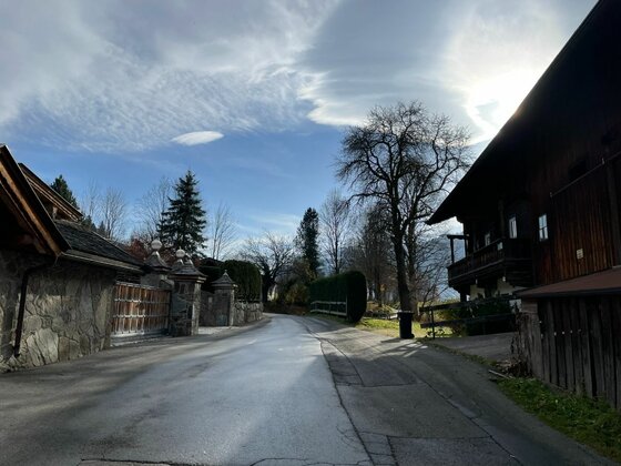 Dorfstraße der Kitzbüheler Runde Asphaltierte Straße zwischen traditionellen Holzhäusern und Steinmauern unter bewölktem Himmel.