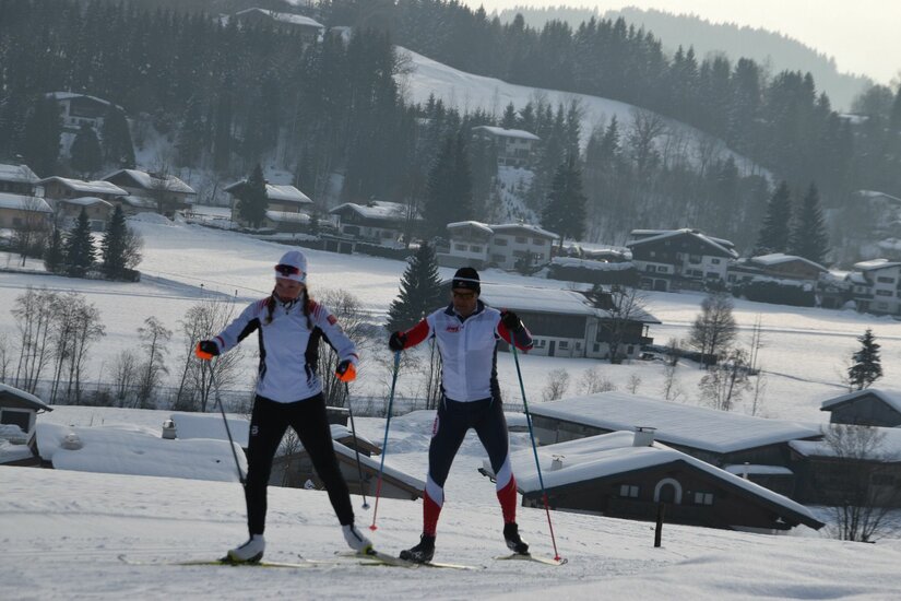 Zwei Personen beim Skilanglauf auf einer verschneiten Loipe vor einer dörflichen Kulisse.
