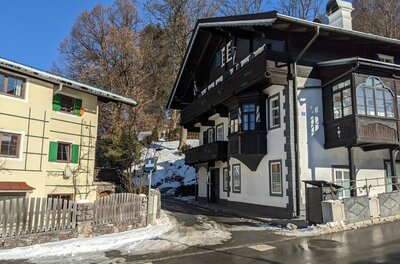 Häuser am Ölberg Zwei mehrstöckige Häuser im alpinen Stil an einer asphaltierten Straße mit Schneeresten unter blauem Himmel.