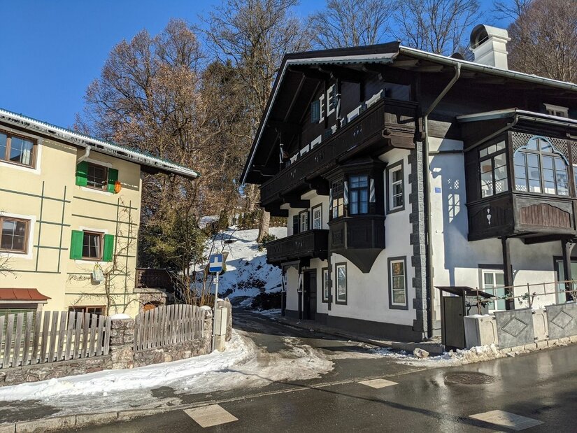 Häuser am Ölberg Zwei mehrstöckige Häuser im alpinen Stil an einer asphaltierten Straße mit Schneeresten unter blauem Himmel.