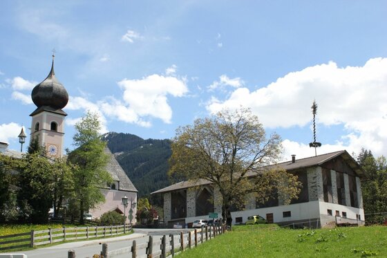 Kirche mit Zwiebelturm und ein größeres Holzgebäude neben einer Straße vor bewaldeten Bergen.
