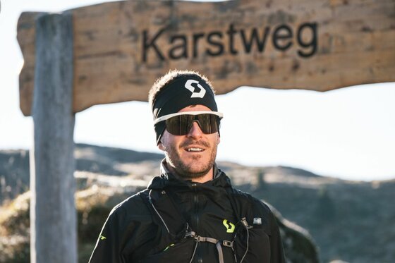 Trailrunner am Karstweg Ein Trailrunner mit Sonnenbrille und Stirnband steht vor einem Holzschild mit der Aufschrift Karstweg.