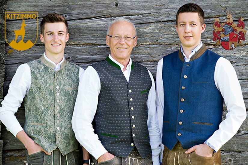 KITZIMMO Immobilientreuhänder Drei Männer stehen vor einer Holzwand und tragen traditionelle Trachtenwesten. Sie lächeln freundlich in die Kamera.